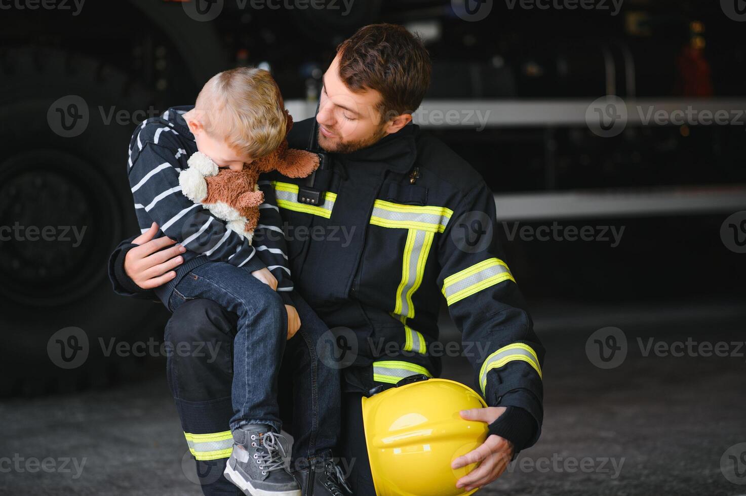 Dirty firefighter in uniform holding little saved boy standing on black background. photo