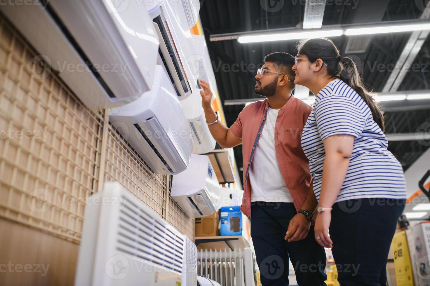 Young indian couple, satisfied customers choosing air conditioner in appliances store photo