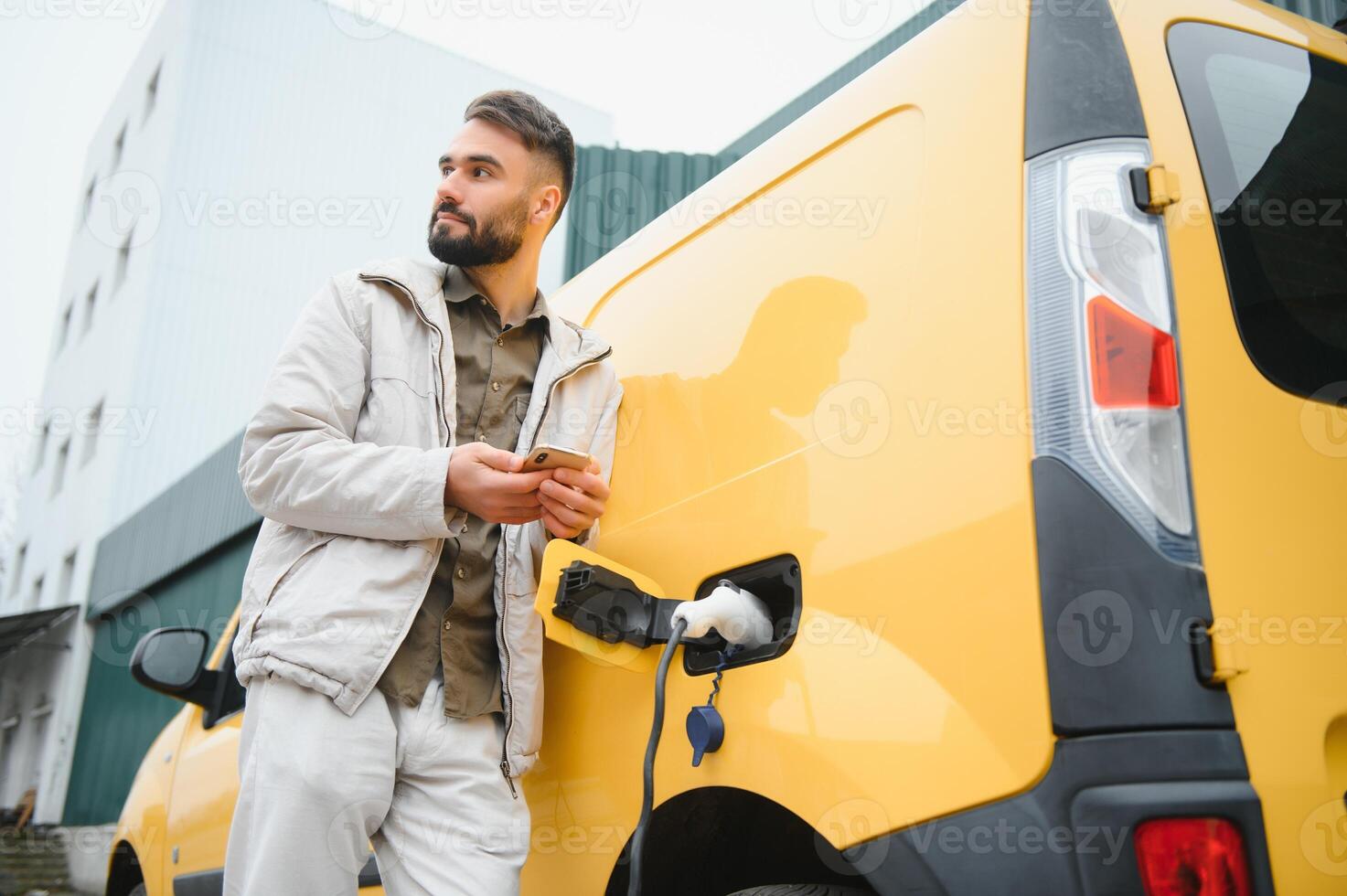 bearded caucasian man standing near an electric car that is charging and making time adjustments on a smartphone. photo