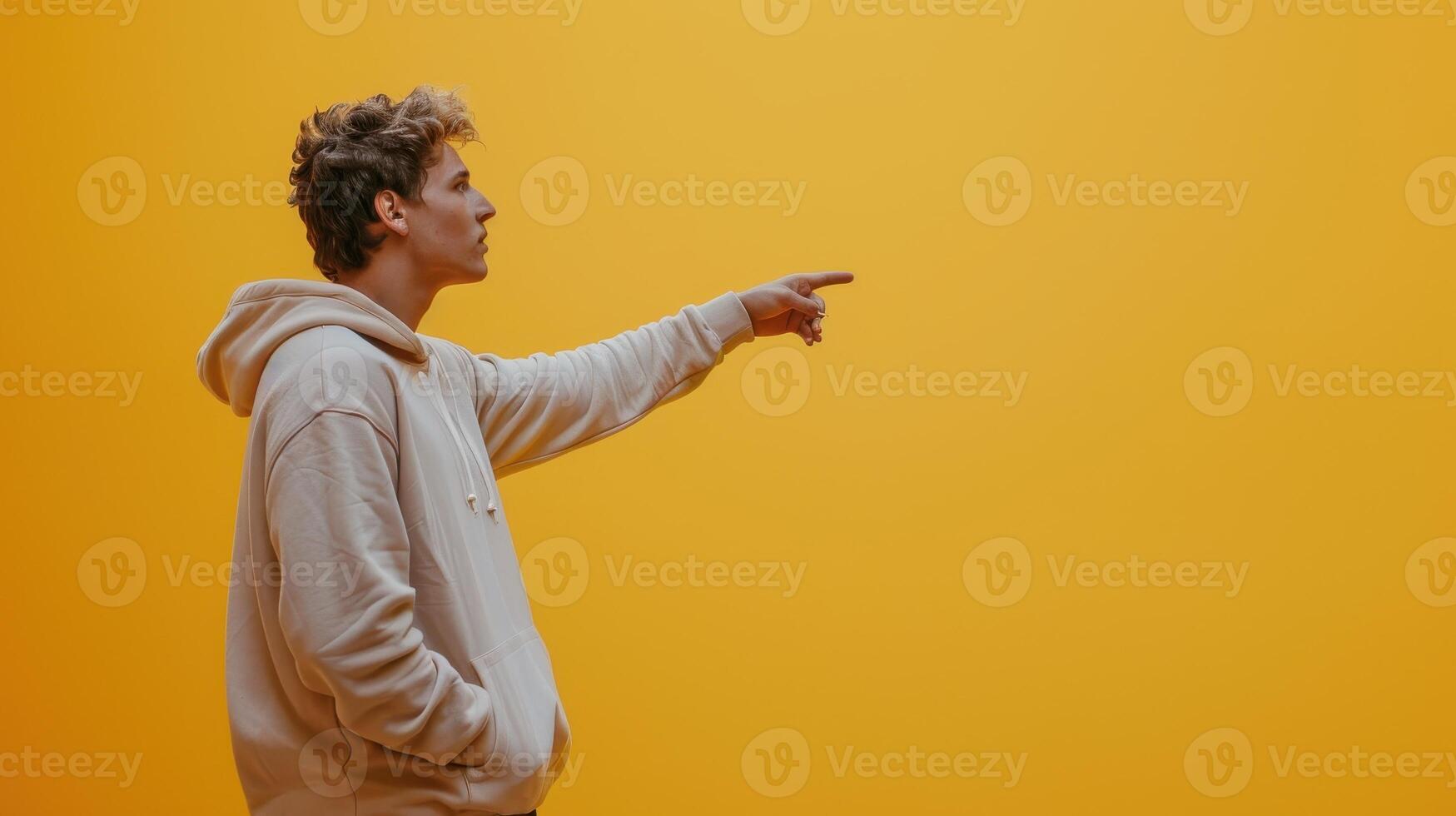 Young Man In A Hooded Sweatshirt Points To The Right In Front Of A Yellow Wall photo