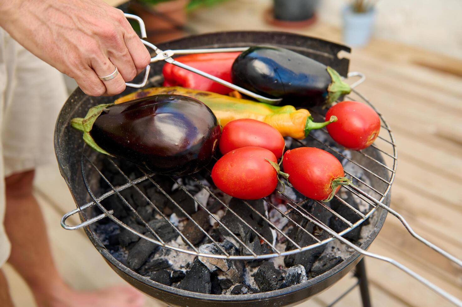 Close-up of a person's hand grilling fresh vegetables, including eggplant, tomatoes and peppers, on a charcoal grill. photo