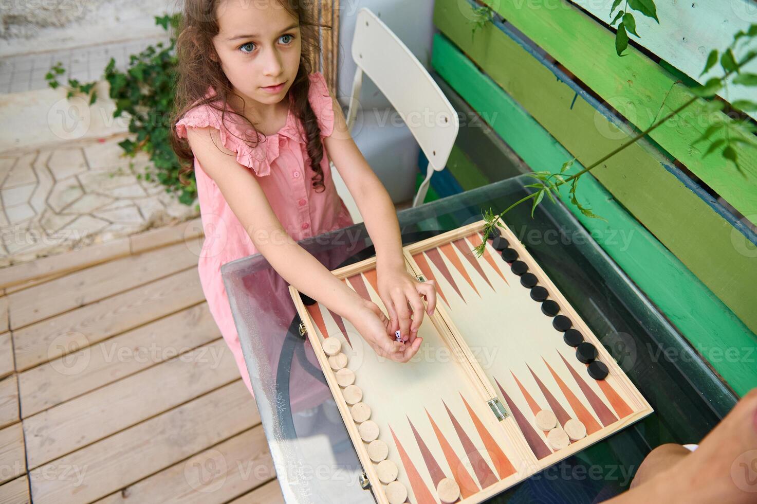Cute girl playing backgammon at an outdoor wooden table in a colorful setting photo