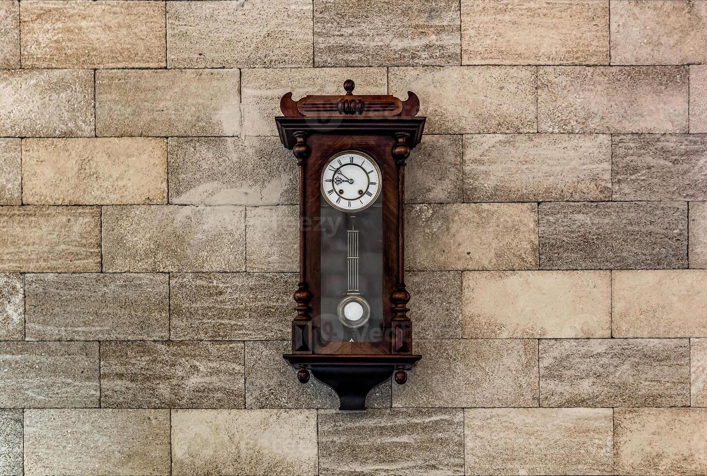 Wall vintage wooden clock with a pendulum on a brick wall. Mechanical watch with Roman numerals on the dial. Light back. photo