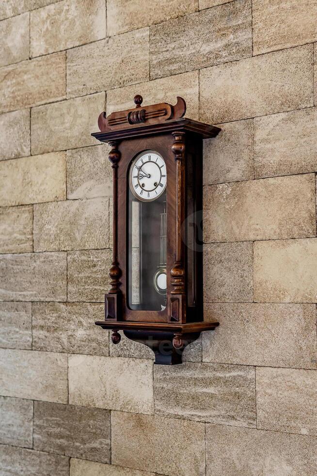 Wall vintage wooden clock with a pendulum on a brick wall. Mechanical watch with Roman numerals on the dial. Light back. photo