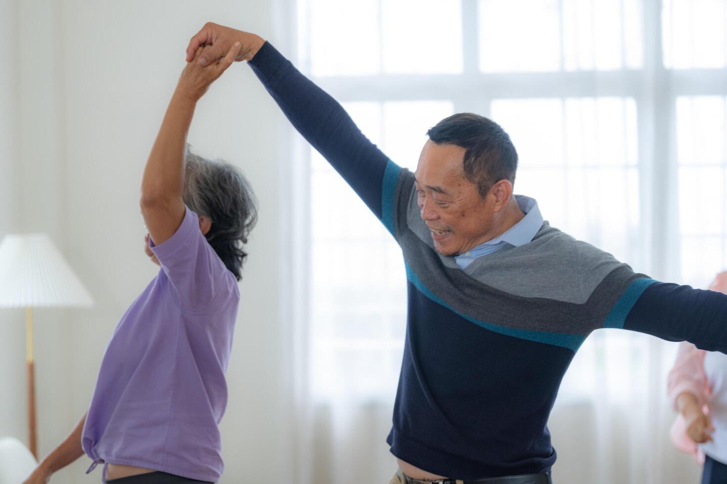 Asian Older male and females people dance with their partners on a dancing floor in living space. Happy older couple performing get exercise. Joyful carefree retired senior friends enjoying relaxation photo