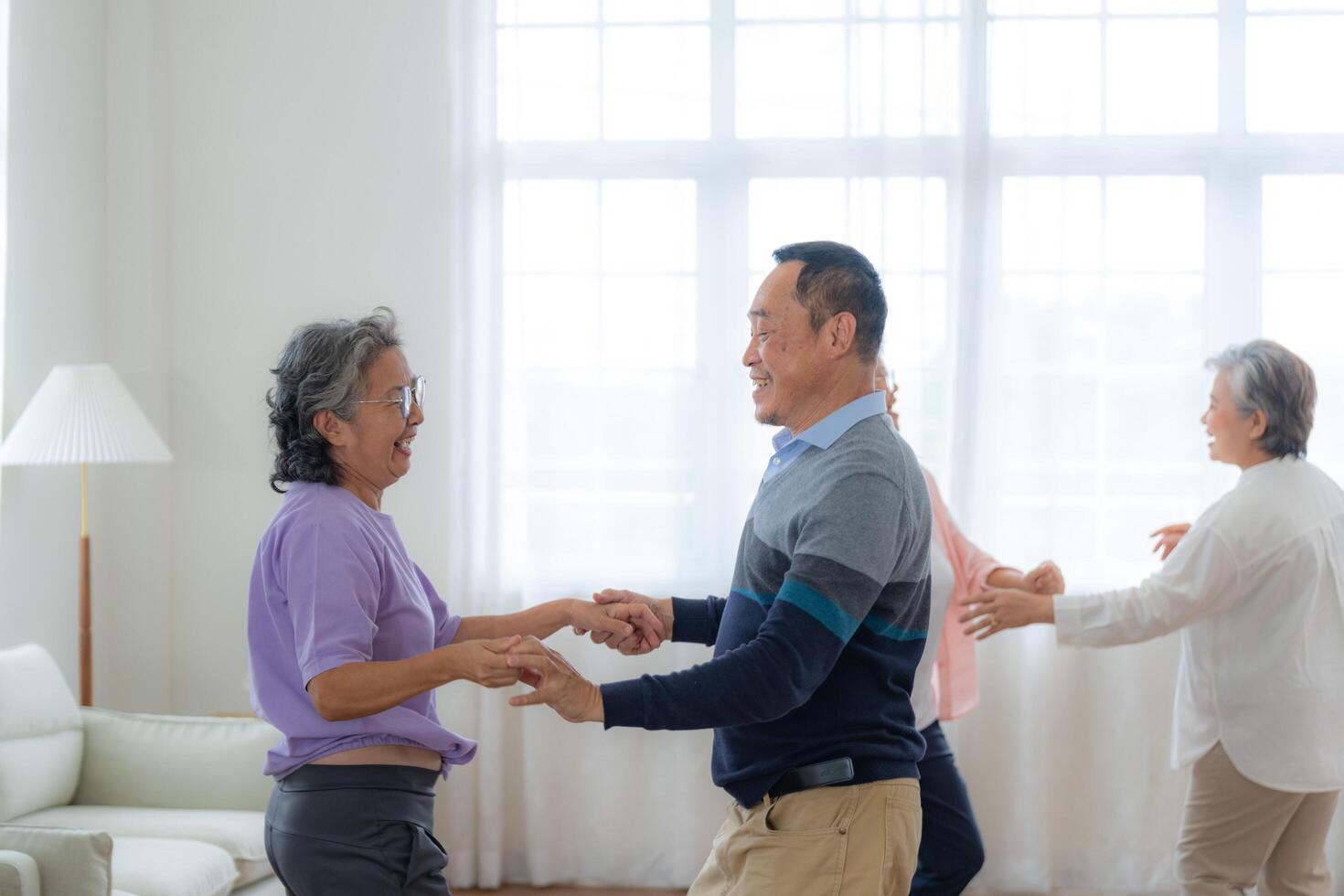 Asian Older male and females people dance with their partners on a dancing floor in living space. Happy older couple performing get exercise. Joyful carefree retired senior friends enjoying relaxation photo