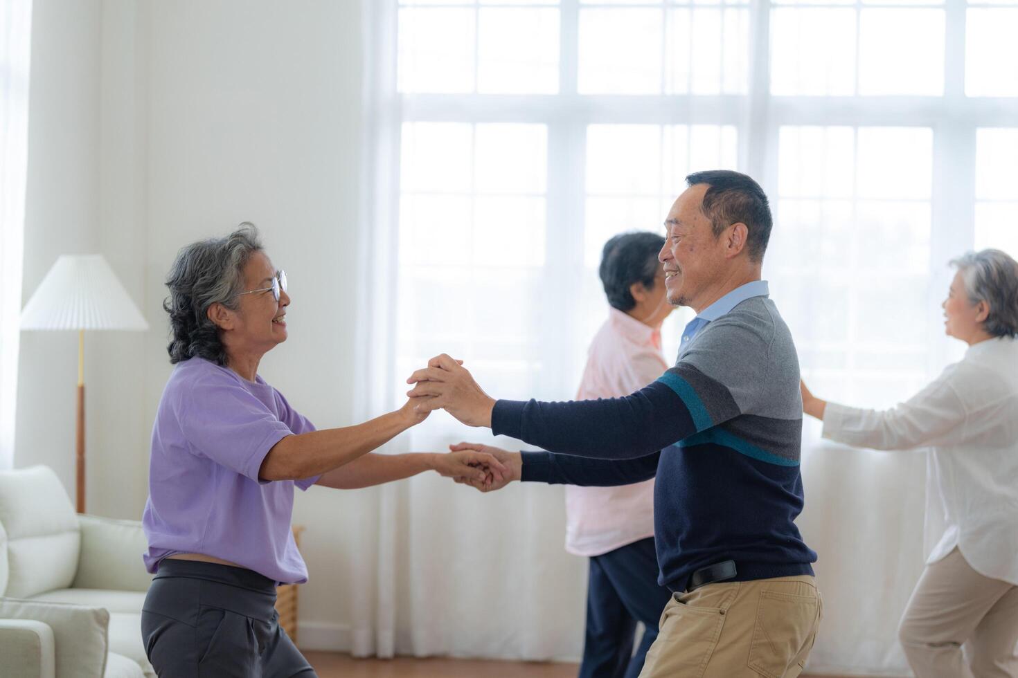 Asian Older male and females people dance with their partners on a dancing floor in living space. Happy older couple performing get exercise. Joyful carefree retired senior friends enjoying relaxation photo