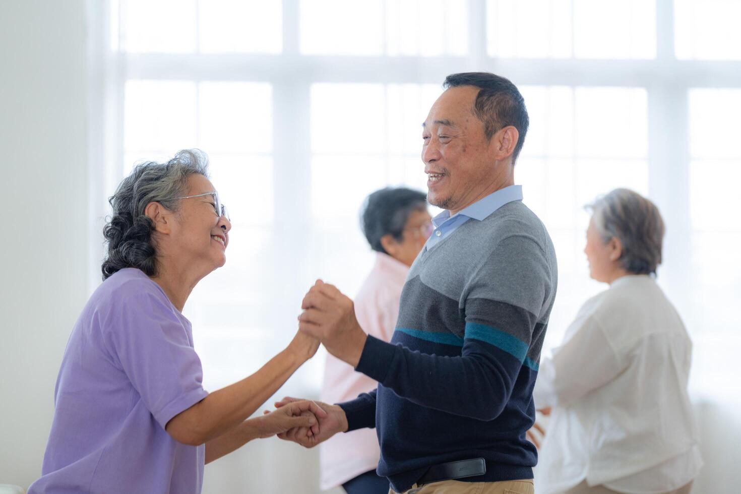 Asian Older male and females people dance with their partners on a dancing floor in living space. Happy older couple performing get exercise. Joyful carefree retired senior friends enjoying relaxation photo