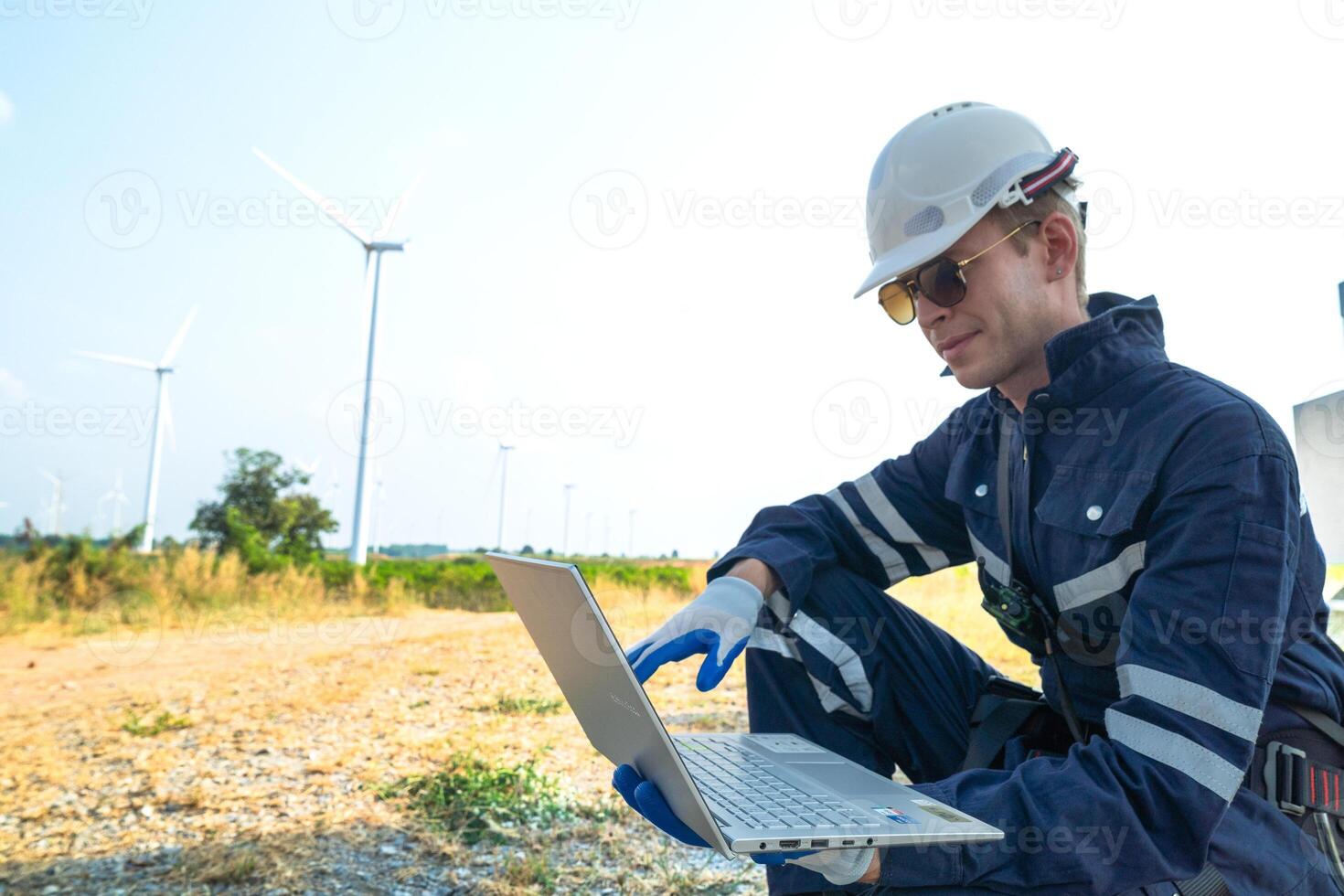 A technician in safety gear meticulously records data with a wind turbine in the backdrop at dusk. photo