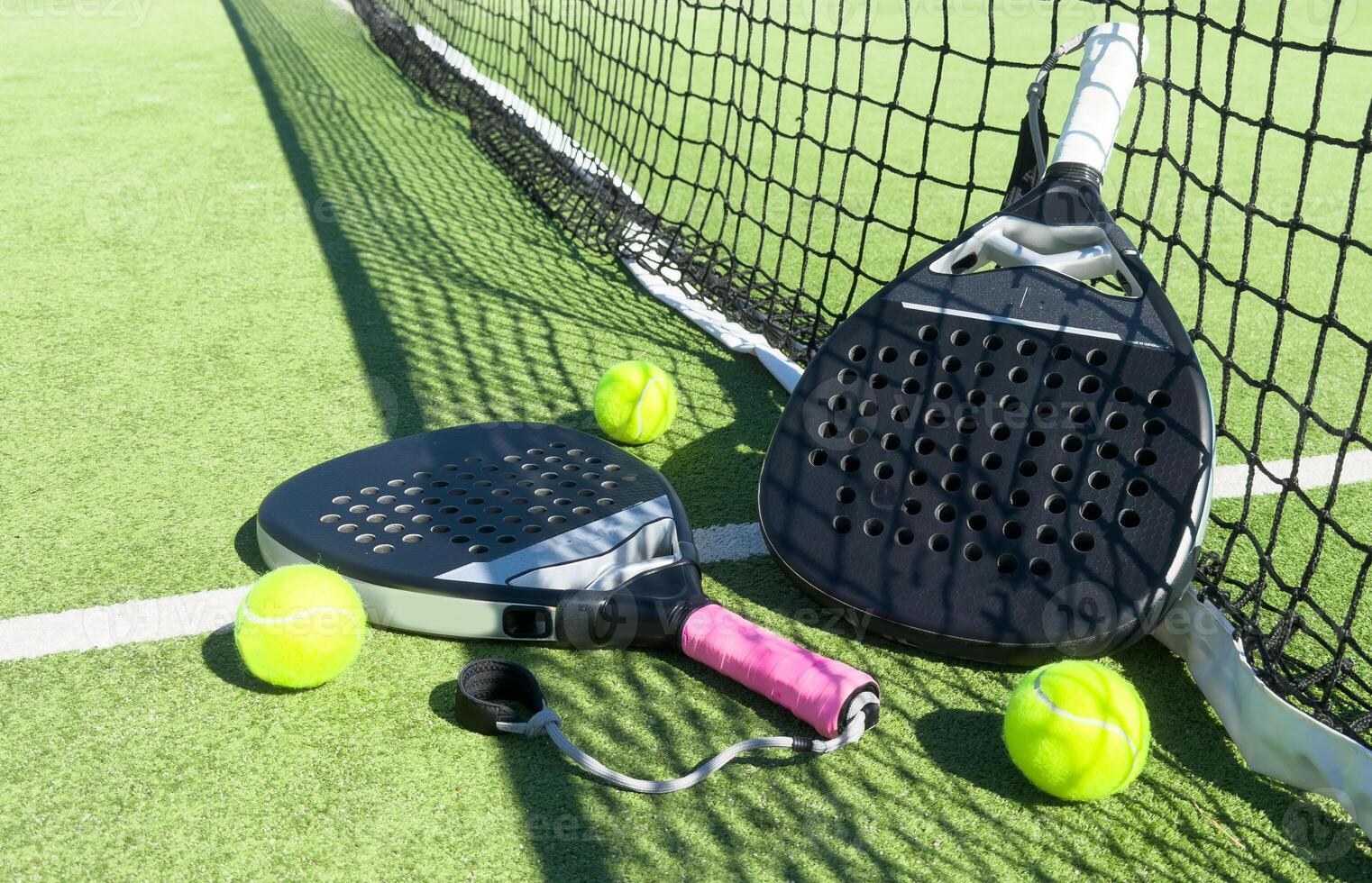 Yellow balls on grass turf near padel tennis racket behind net in green court outdoors with natural lighting. Paddle is a racquet game. Professional sport concept with copy space. photo