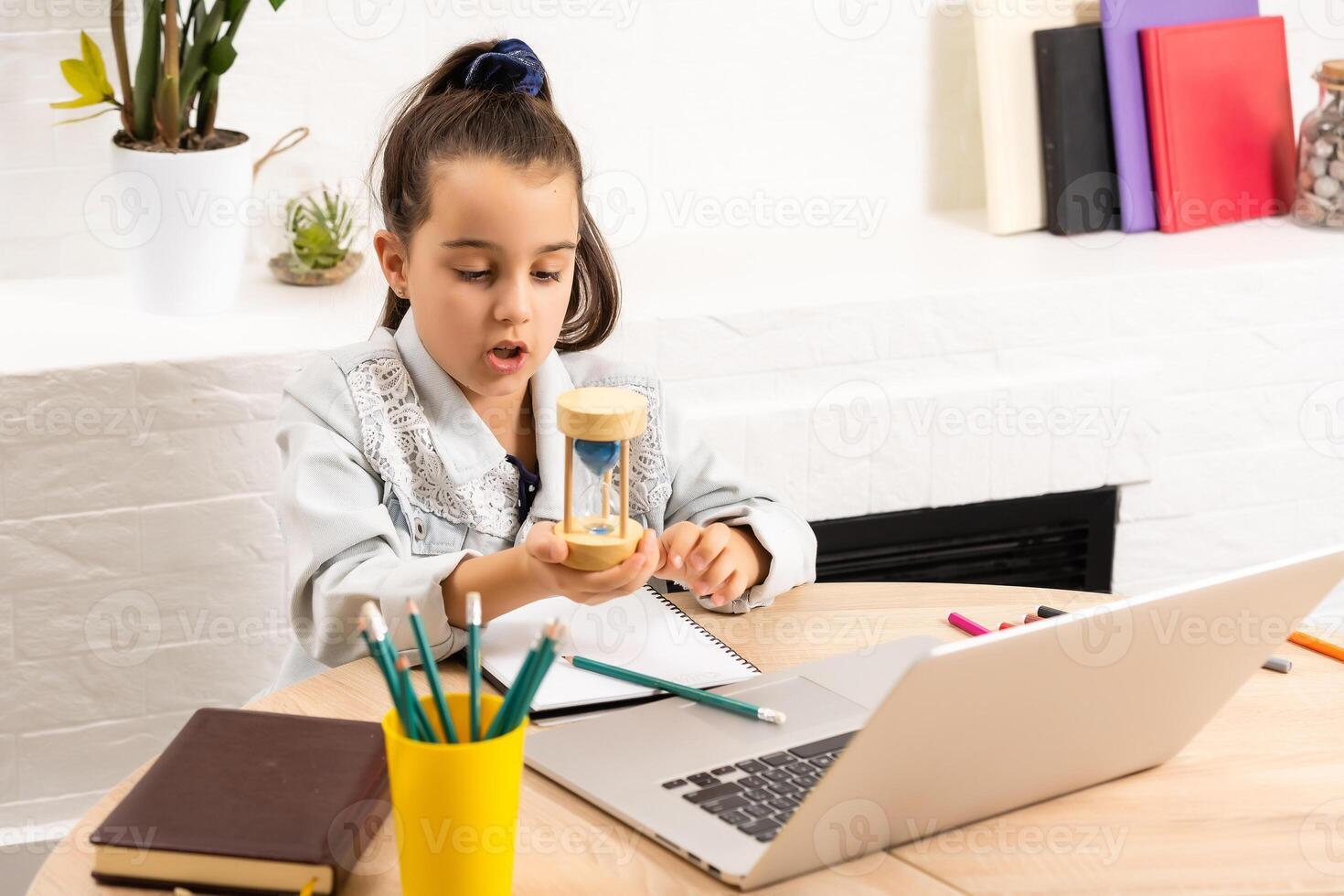 little girl sits with a laptop at the table and holds an hourglass, waiting for the end of online lessons photo