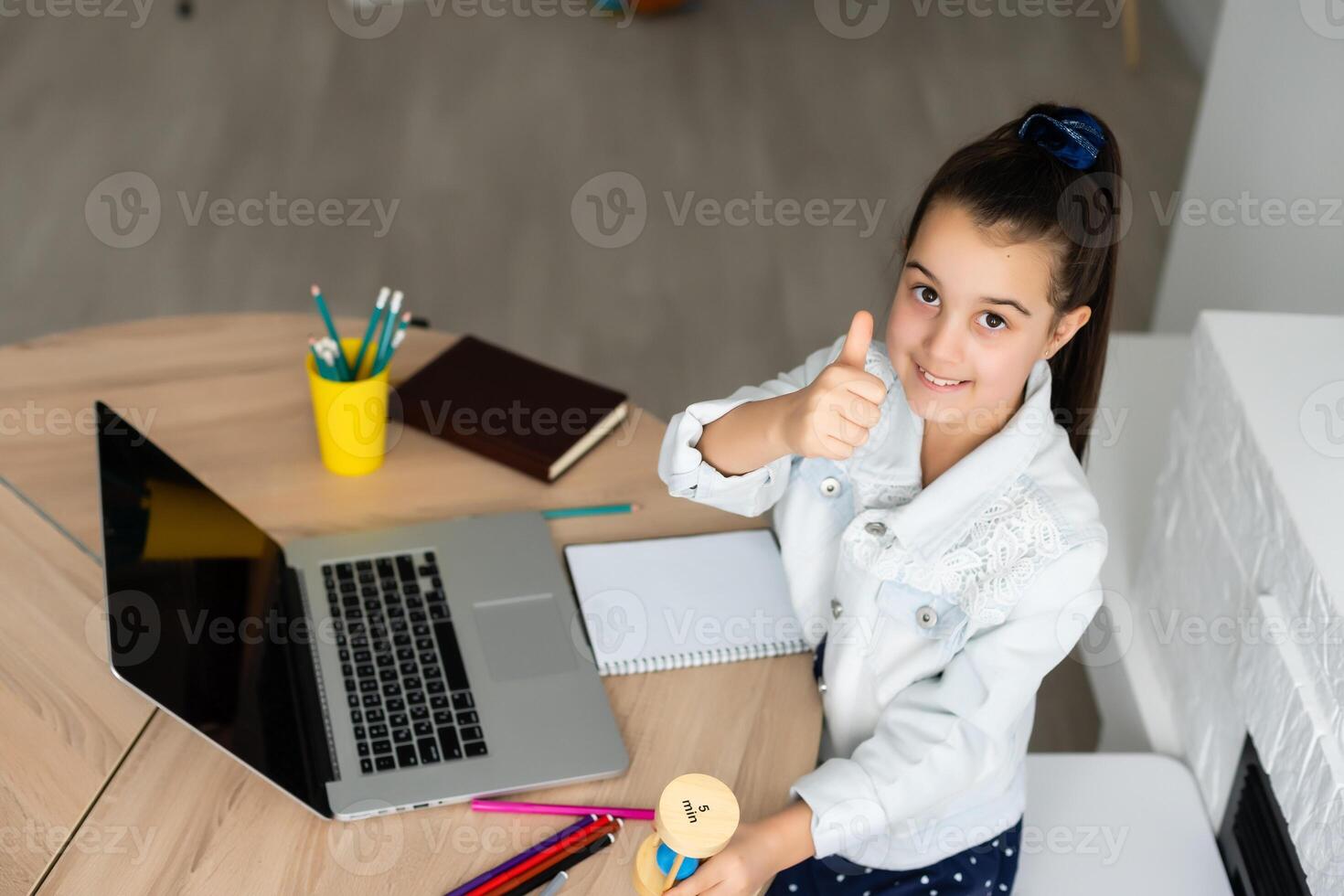 little girl sits with a laptop at the table and holds an hourglass, waiting for the end of online lessons photo