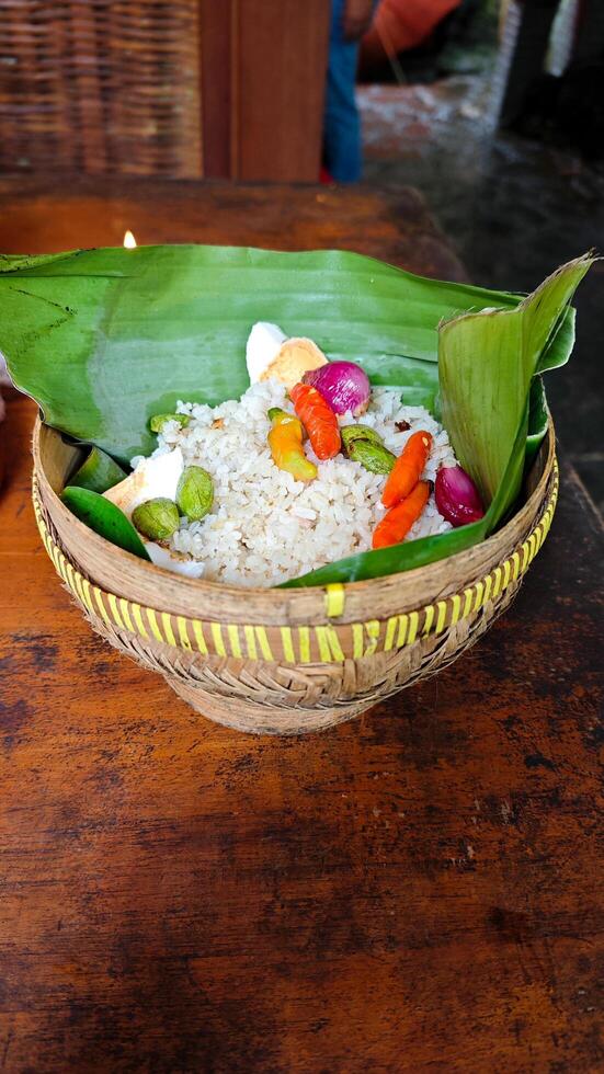 Traditional Indonesian Nasi Bakul in Woven Bamboo Basket with Spicy Garnish and Banana Leaf on Rustic Wooden Table photo