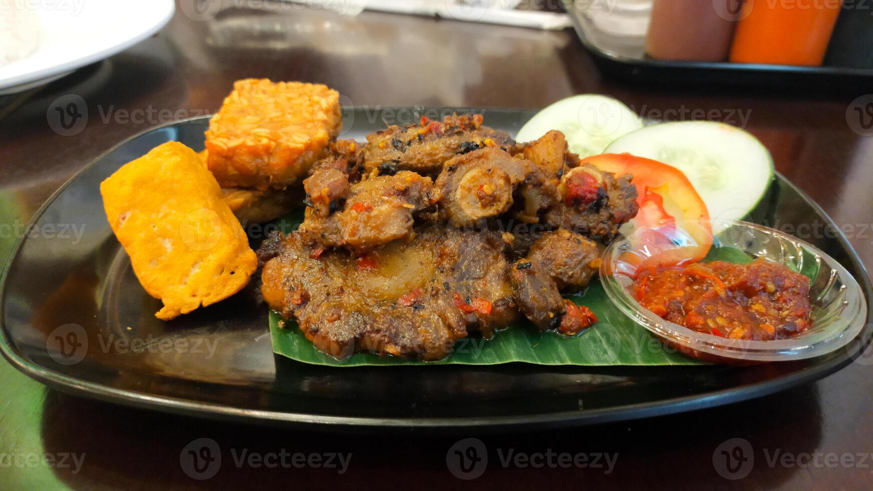 Spicy Grilled Oxtail with Tofu and Tempeh Served with Fresh Cucumber and Tomato Slices and Spicy Chili Sauce on Black Plate photo