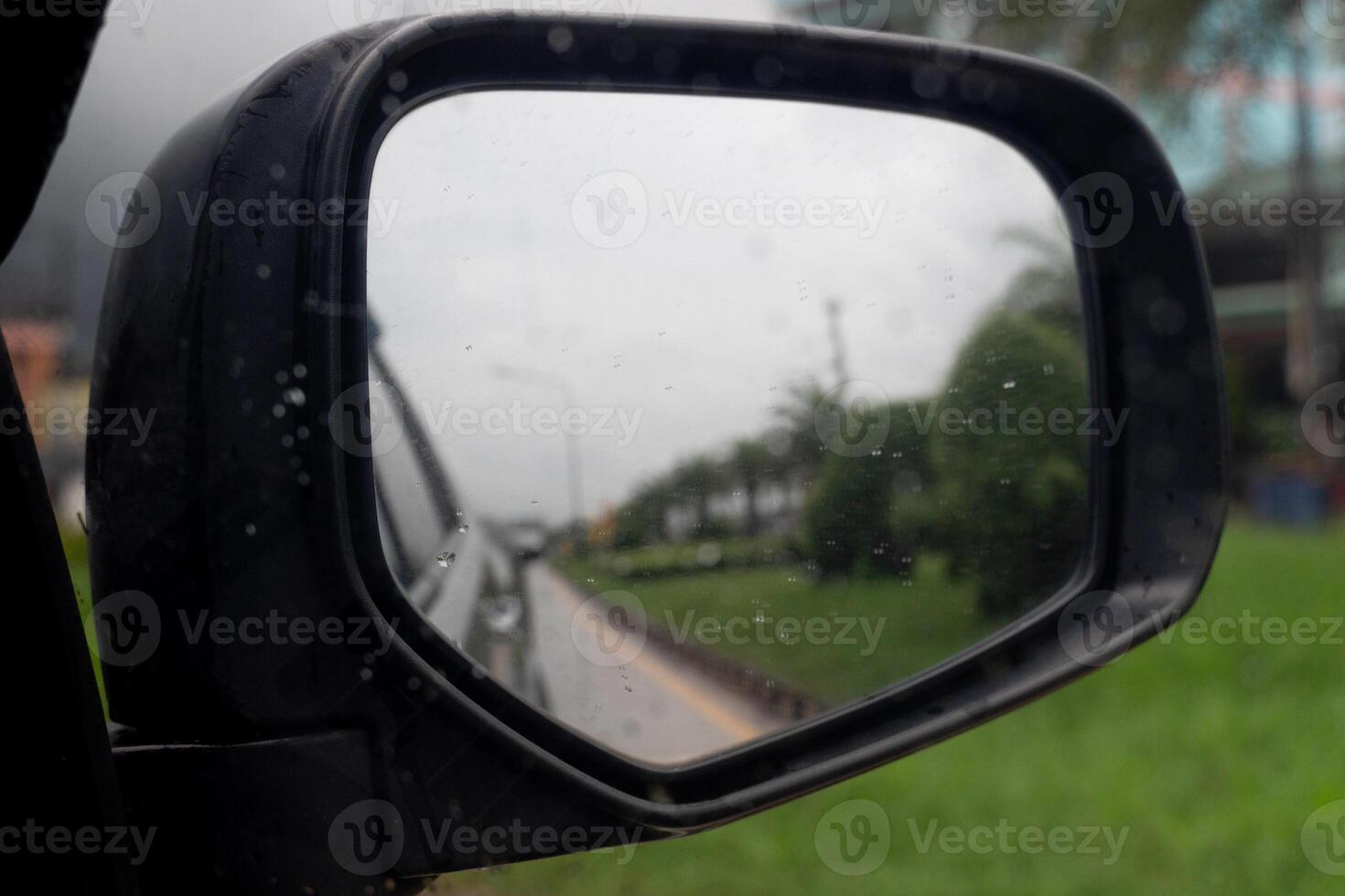 Inside view of mirrors wing rear view of a gray car with drop of water rain on the glass. Driving on aspahlt road beside with green grass. Under of white clouds of rainy. photo