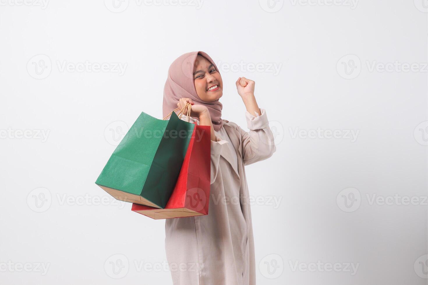 Portrait of excited Asian hijab woman in casual suit holding paper shopping bags while raising her fist. Shopaholic girl and discount buying concept. Isolated image on white background photo