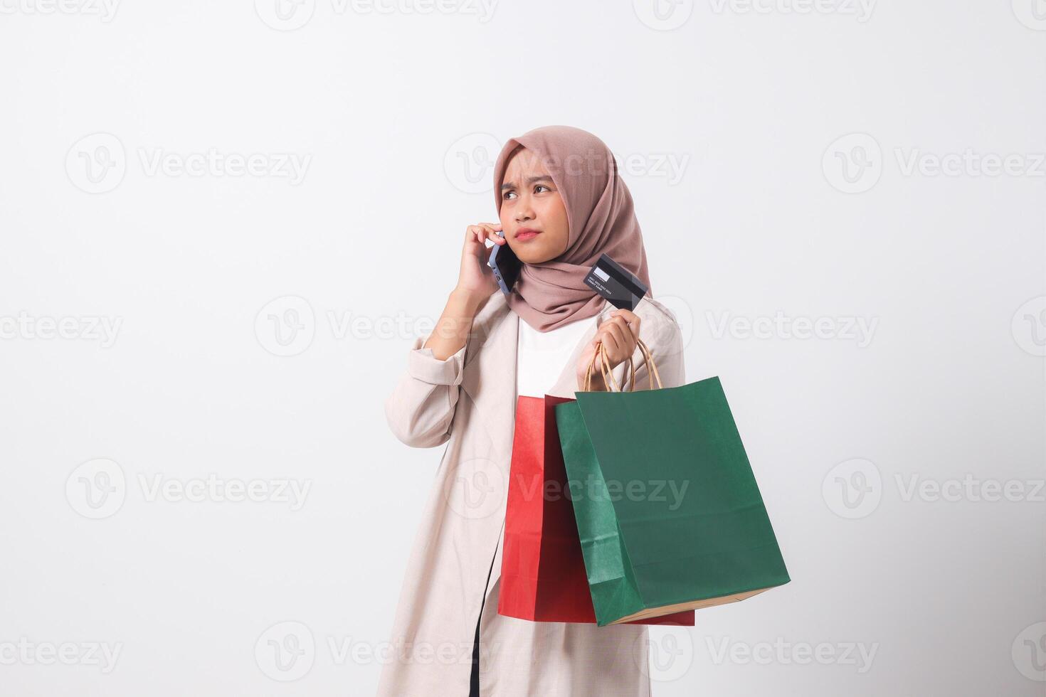 Portrait of depressed Asian hijab woman in casual suit holding credit card and shopping bags while using mobile phone. Shopaholic girl and discount buying concept. Isolated image on white background photo