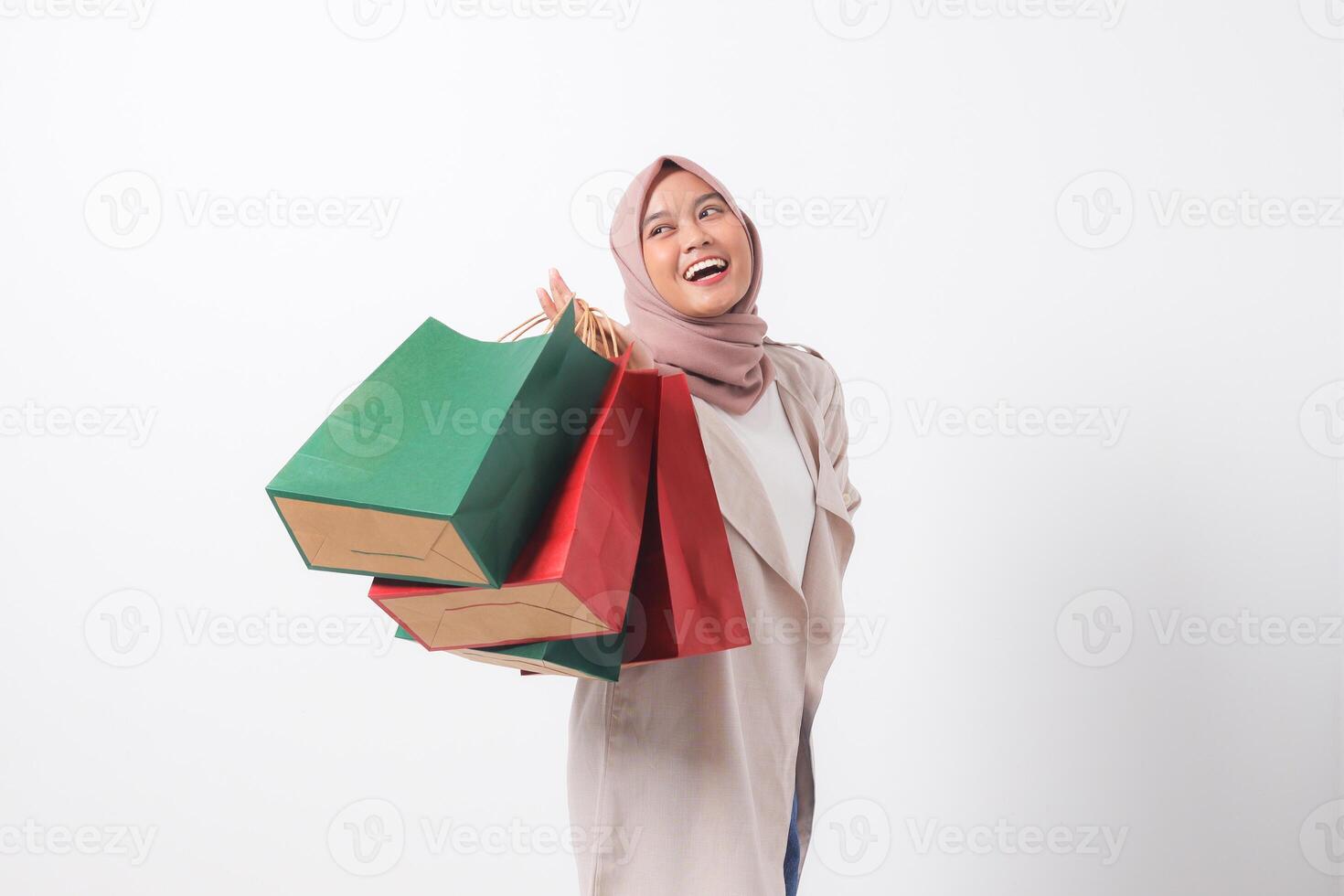 Portrait of excited Asian hijab woman in casual suit holding and showing colorful paper shopping bags. Shopaholic girl and discount buying concept. Isolated image on white background photo