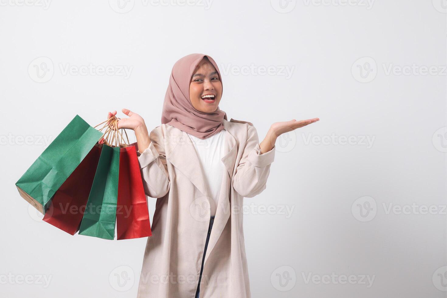 Portrait of excited Asian hijab woman in casual suit holding paper shopping bags while pointing with finger. Shopaholic girl and discount buying concept. Isolated image on white background photo