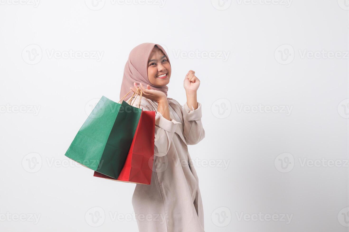 Portrait of excited Asian hijab woman in casual suit holding paper shopping bags while raising her fist. Shopaholic girl and discount buying concept. Isolated image on white background photo