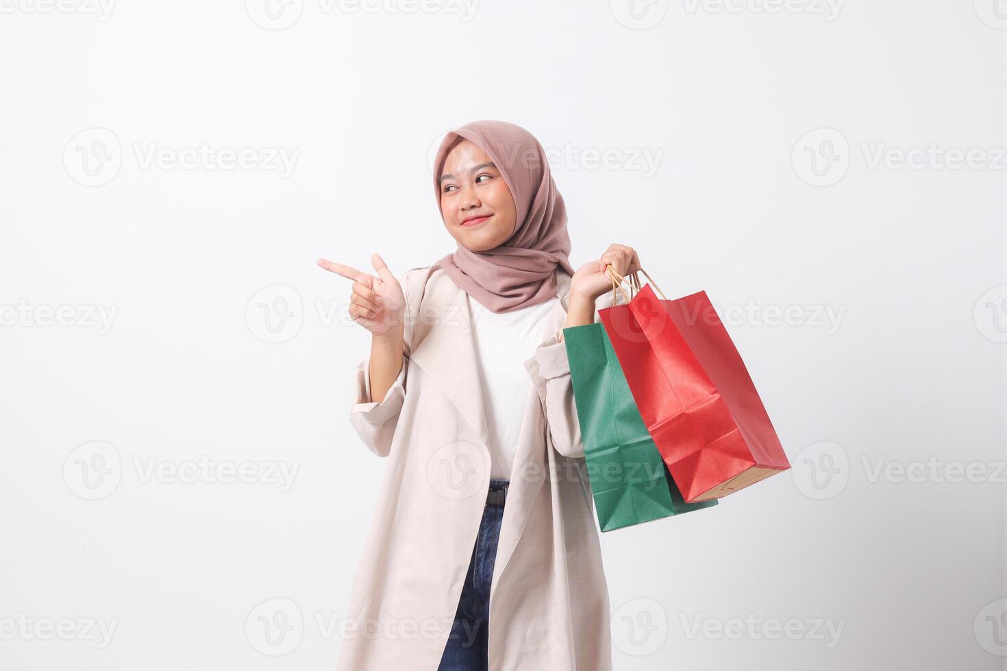 Portrait of excited Asian hijab woman in casual suit holding paper shopping bags while pointing with finger. Shopaholic girl and discount buying concept. Isolated image on white background photo