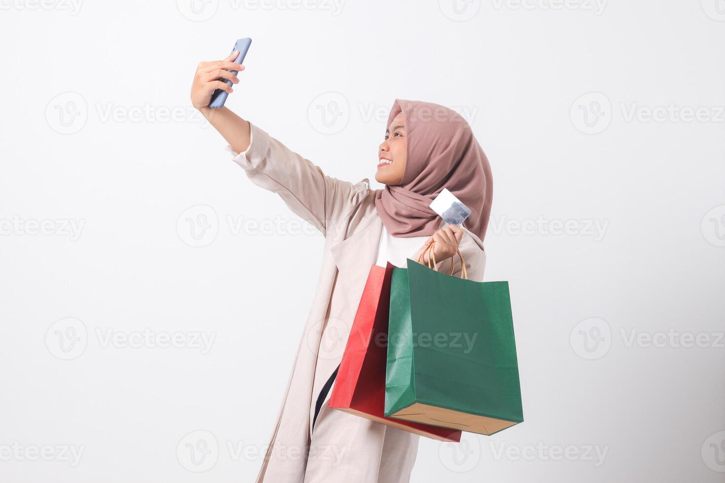 Portrait of excited Asian hijab woman in casual suit holding credit card and shopping bags while taking selfie with mobile phone. Shopaholic girl concept. Isolated image on white background photo