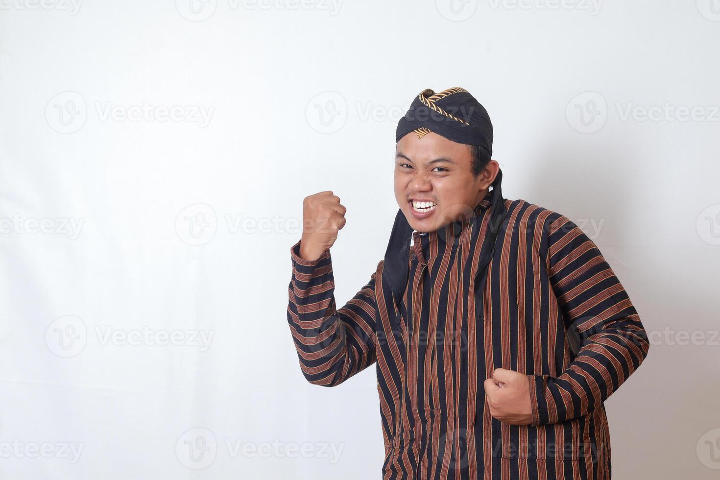 Portrait of excited Asian man in javanese cloth with blangkon hat raising his fist, celebrating success after winning a competition. Isolated image on white background photo