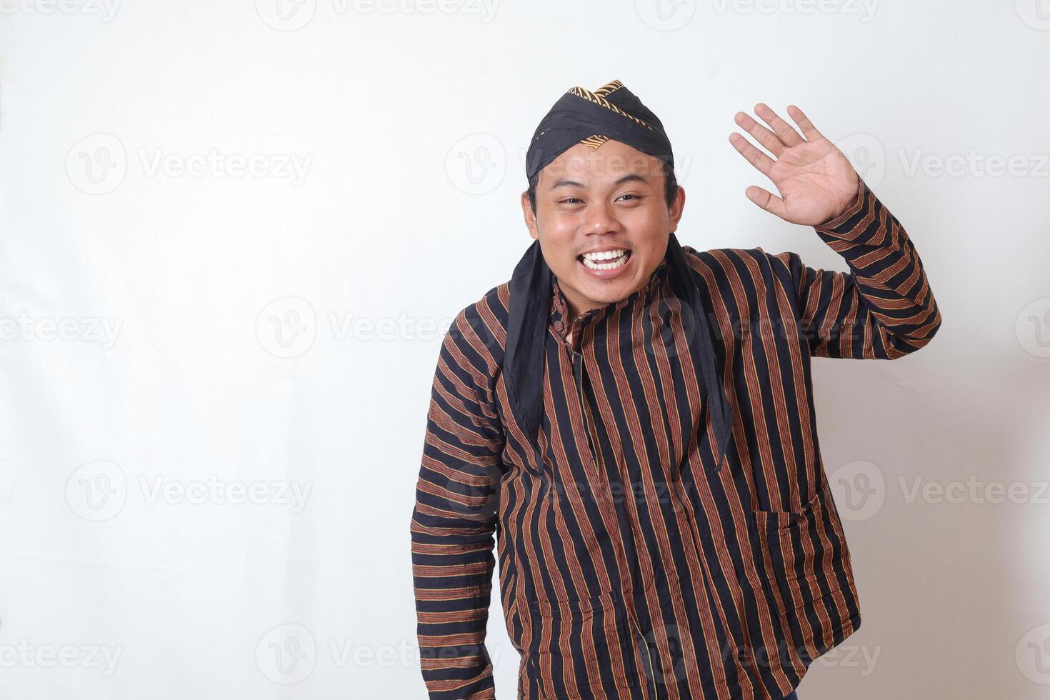 Portrait of friendly Asian man in javanese cloth with blangkon hat saying hi, waving hand at camera, greeting person with happy smile. Isolated image on white background photo