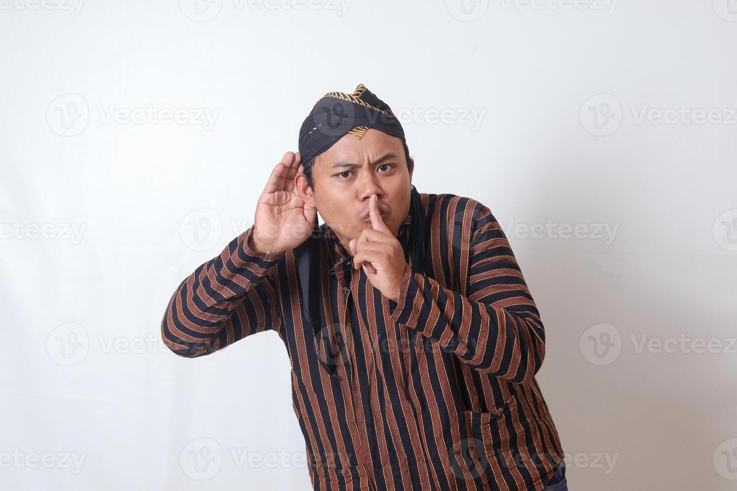 Portrait of serious Asian man in javanese cloth with blangkon hat whispering malicious talk conversation, hand on mouth telling secret rumor. Isolated image on white background photo