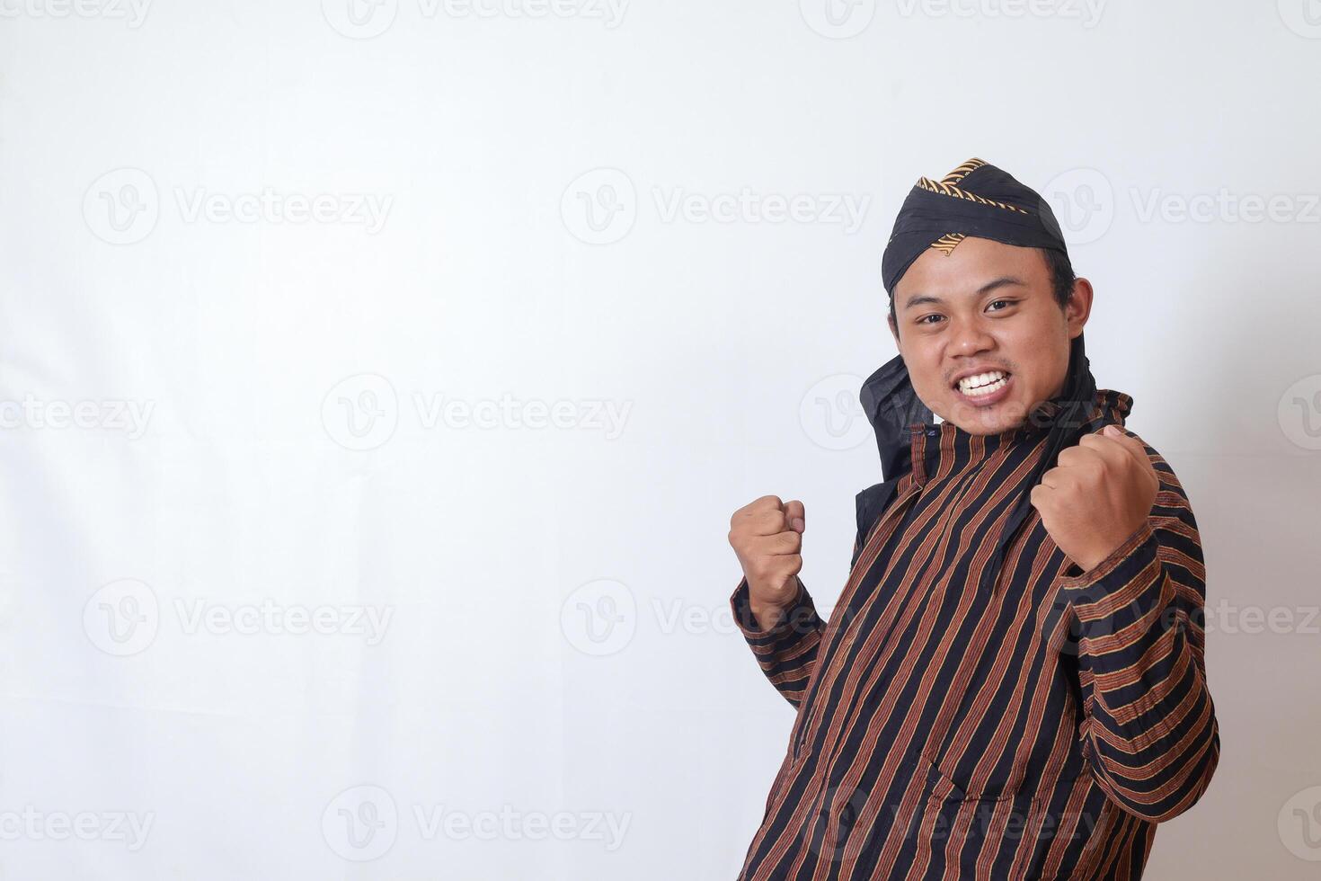 Portrait of excited Asian man in javanese cloth with blangkon hat raising his fist, celebrating success after winning a competition. Isolated image on white background photo