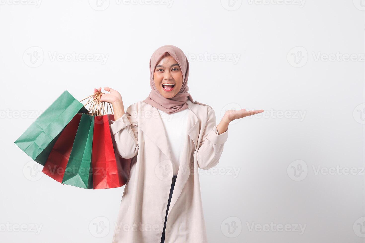 Portrait of excited Asian hijab woman in casual suit holding paper shopping bags while pointing with finger. Shopaholic girl and discount buying concept. Isolated image on white background photo