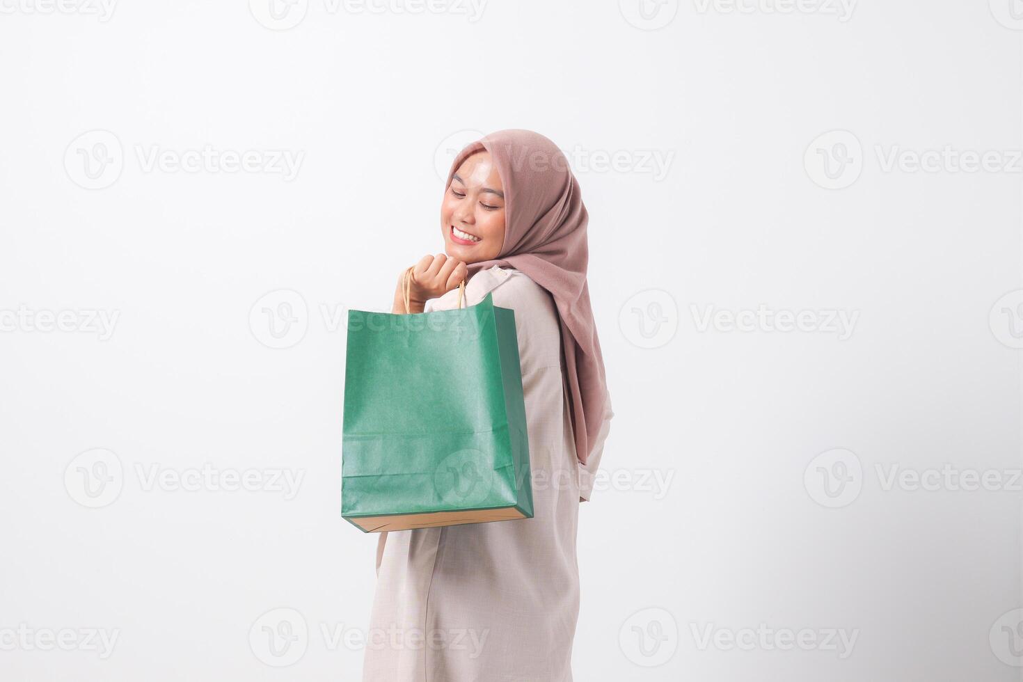 Portrait of cheerful Asian hijab woman in casual suit holding and showing colorful paper shopping bags. Shopaholic girl and discount buying concept. Isolated image on white background photo