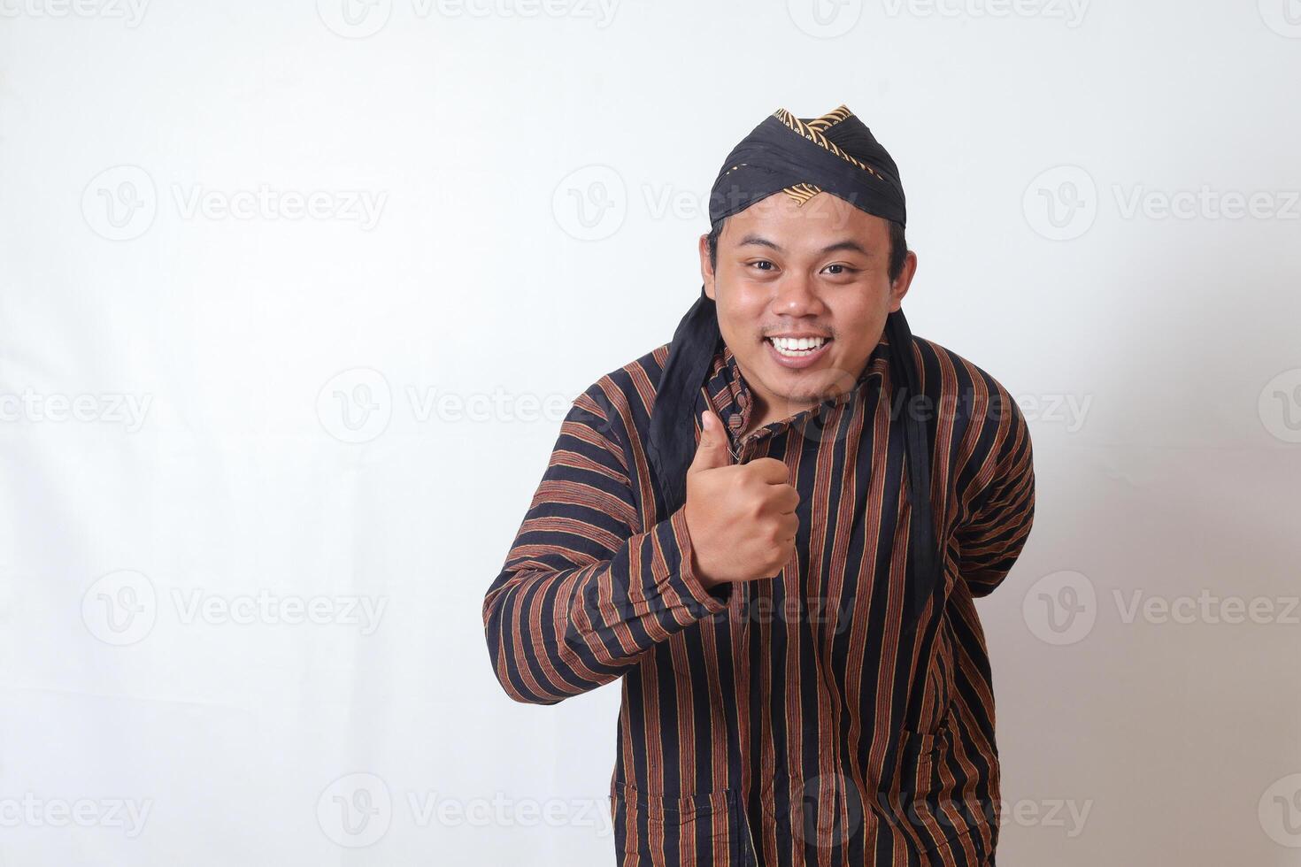 Portrait of amazed Asian man in javanese cloth with blangkon hat showing approval and agreement sign, making thumbs up hand gesture. Isolated image on white background photo