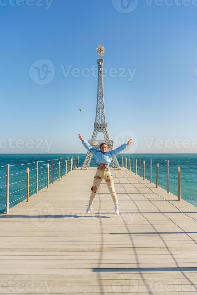 Large model of the Eiffel Tower on the beach. A woman walks along the pier towards the tower, wearing a blue jacket and white jeans. photo