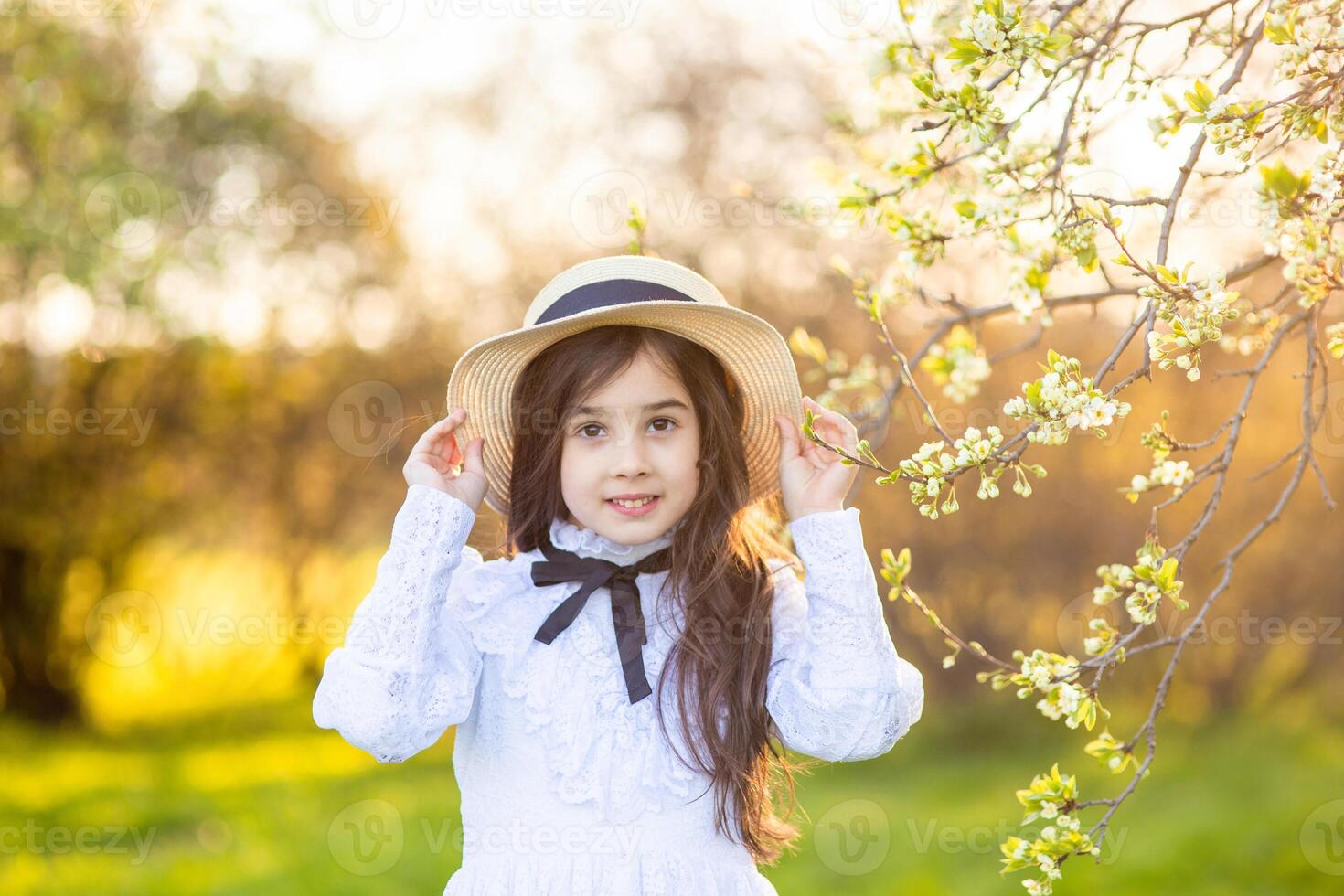 Portrait of cute little girl in a white dress and hat, standing under flowering trees, in spring photo