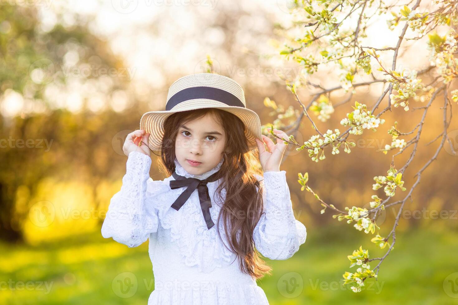 Portrait of beautiful little girl in hat and white dress , standing under flowering trees, in spring photo