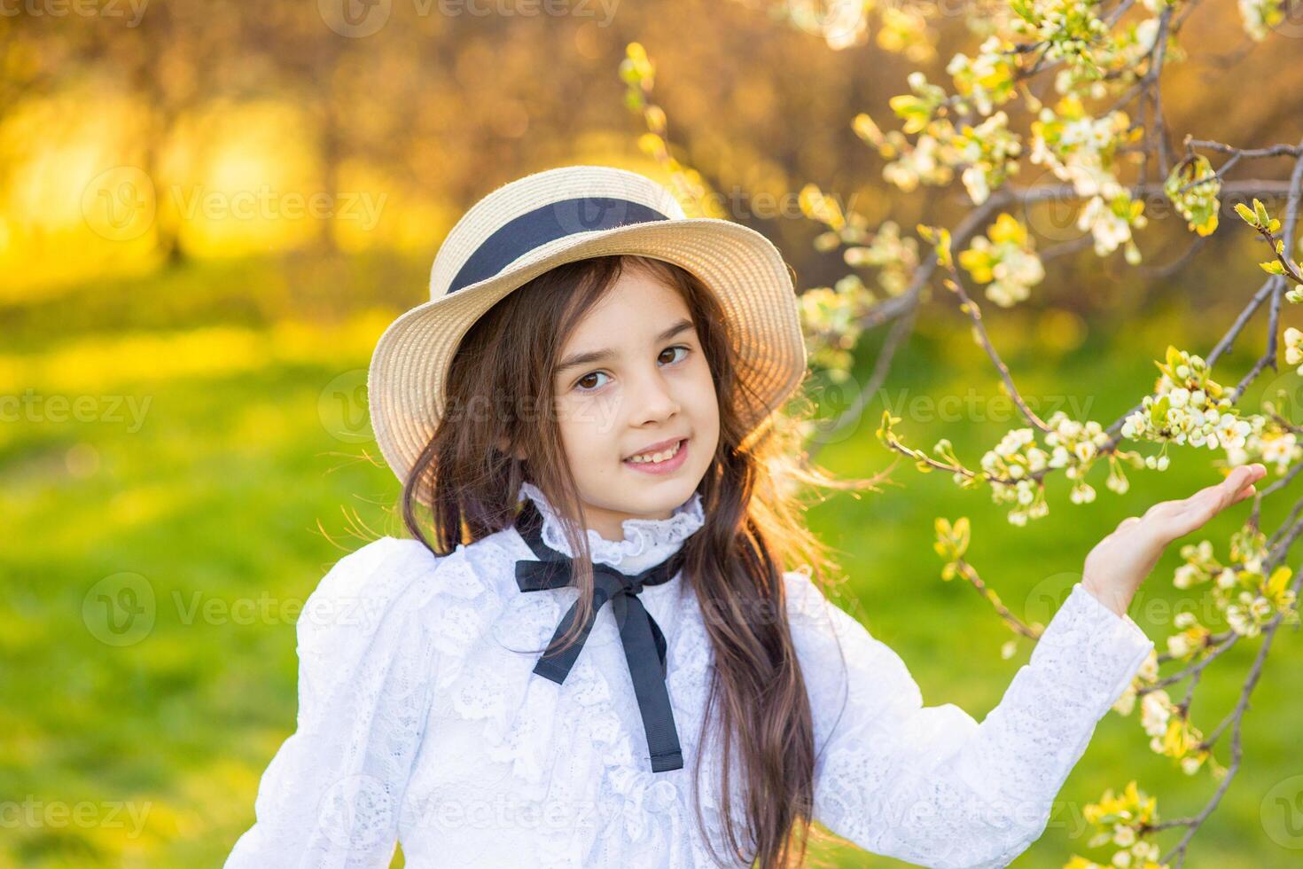 a adorable girl in a hat and white openwork dress, holding a branch of a tree blooming with white flowers photo