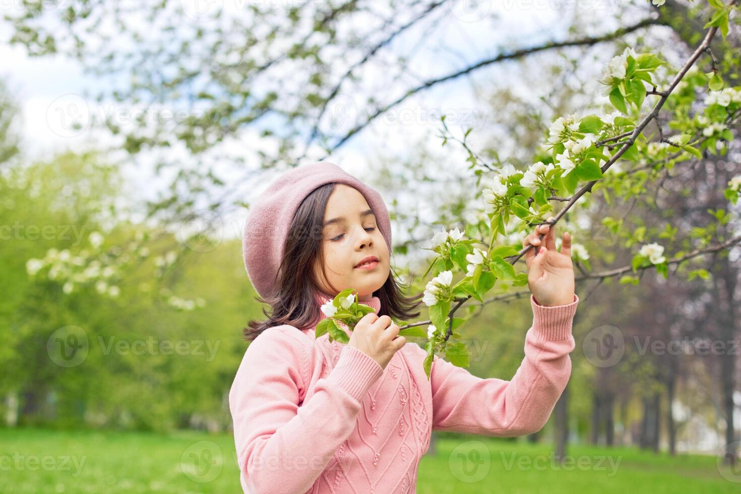 A beautiful girl in a pink jumper holds a branch of a blossoming white apple tree in the park during the day photo