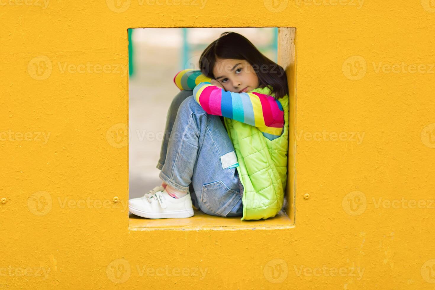 Sad and bored cute little girl is sitting among yellow colorful playground in summer. photo