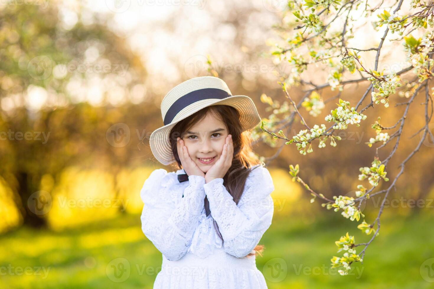 Portrait of adorable little girl in a white dress and hat, standing under flowering trees, in spring photo