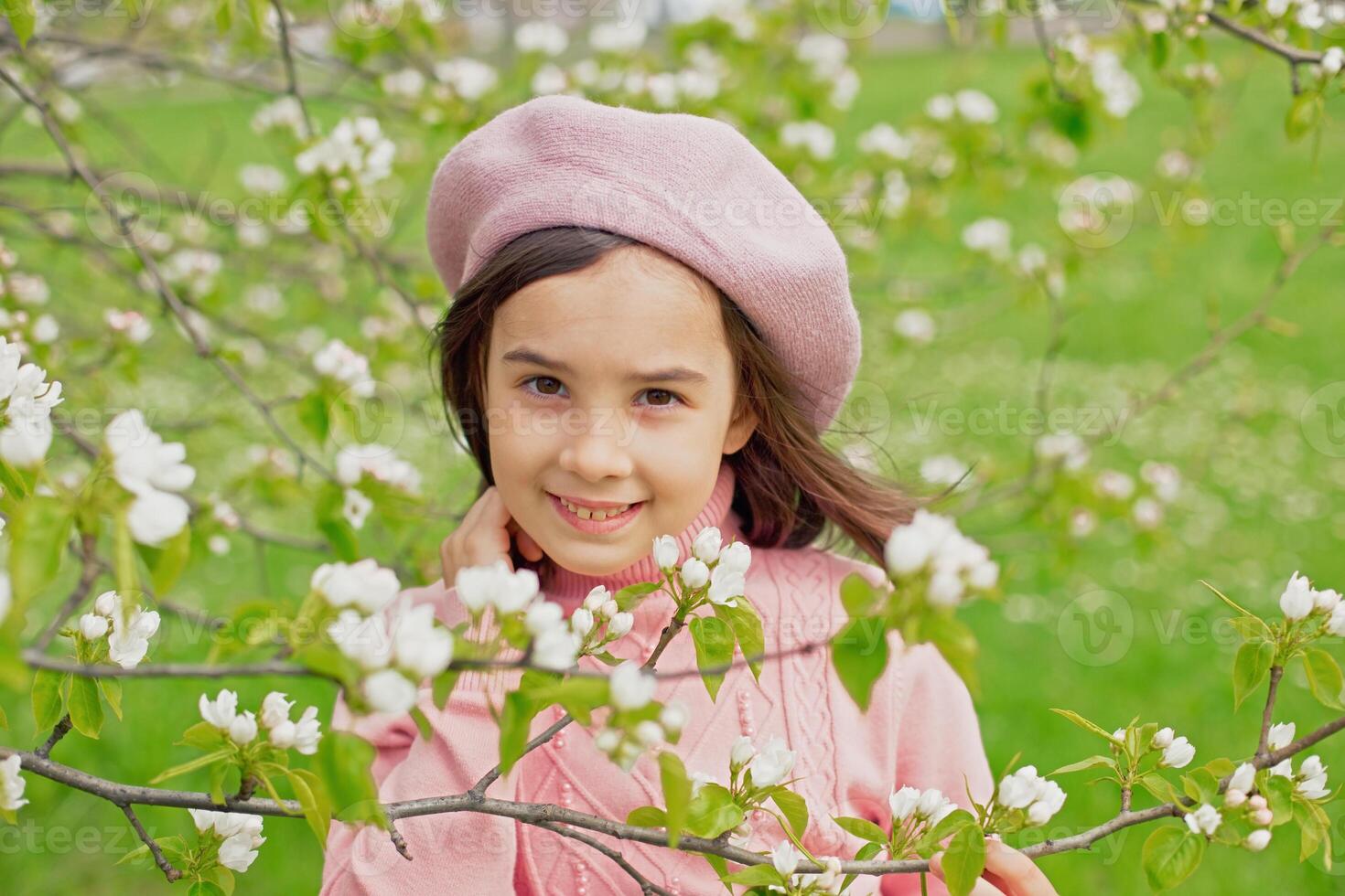 Portrait of a beautiful little girl stands next to the branches of a blossoming white apple tree in the park. photo