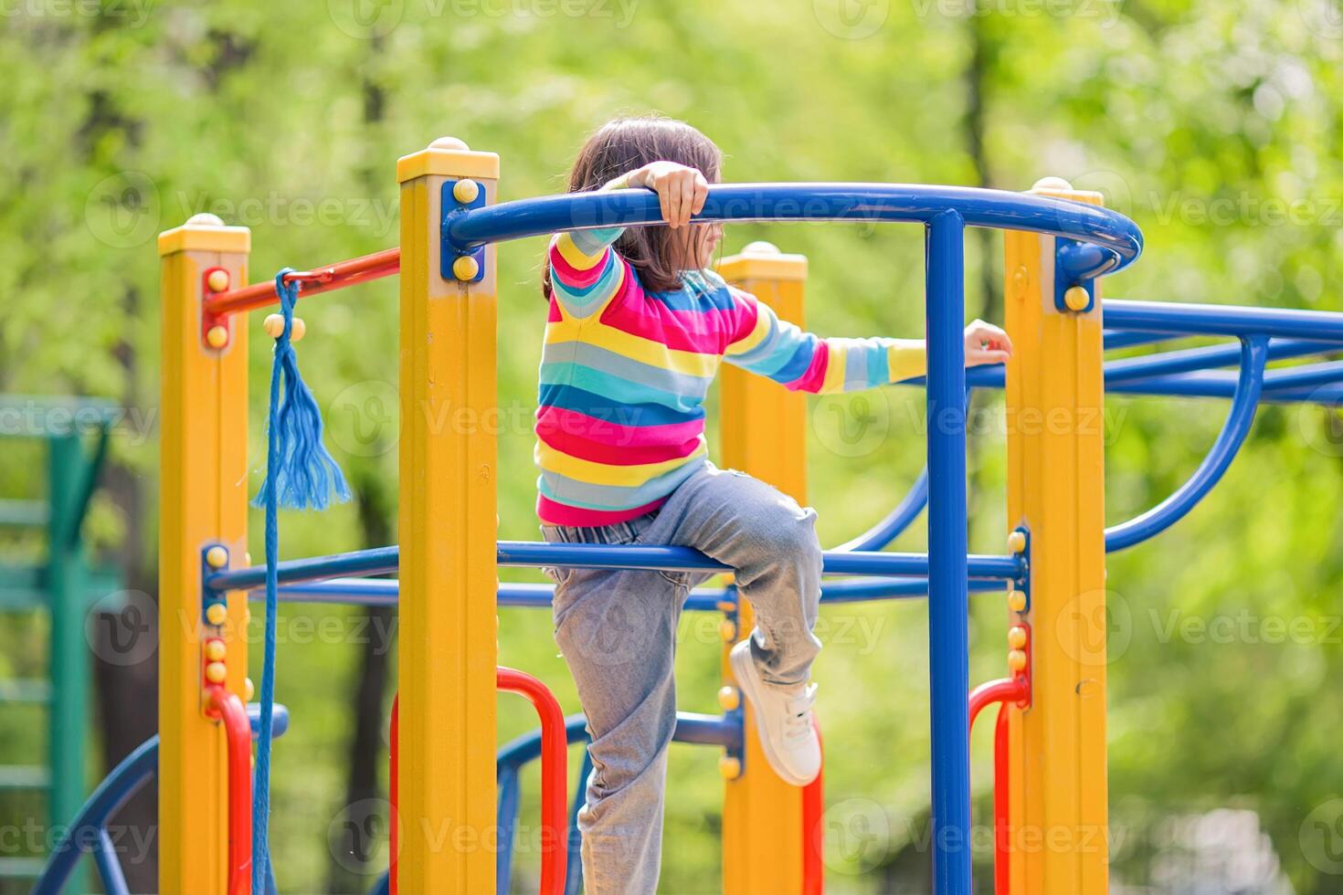 A little girl 5-6 years old, in a bright striped jumper, climbs on a multi-colored playground, in a park photo