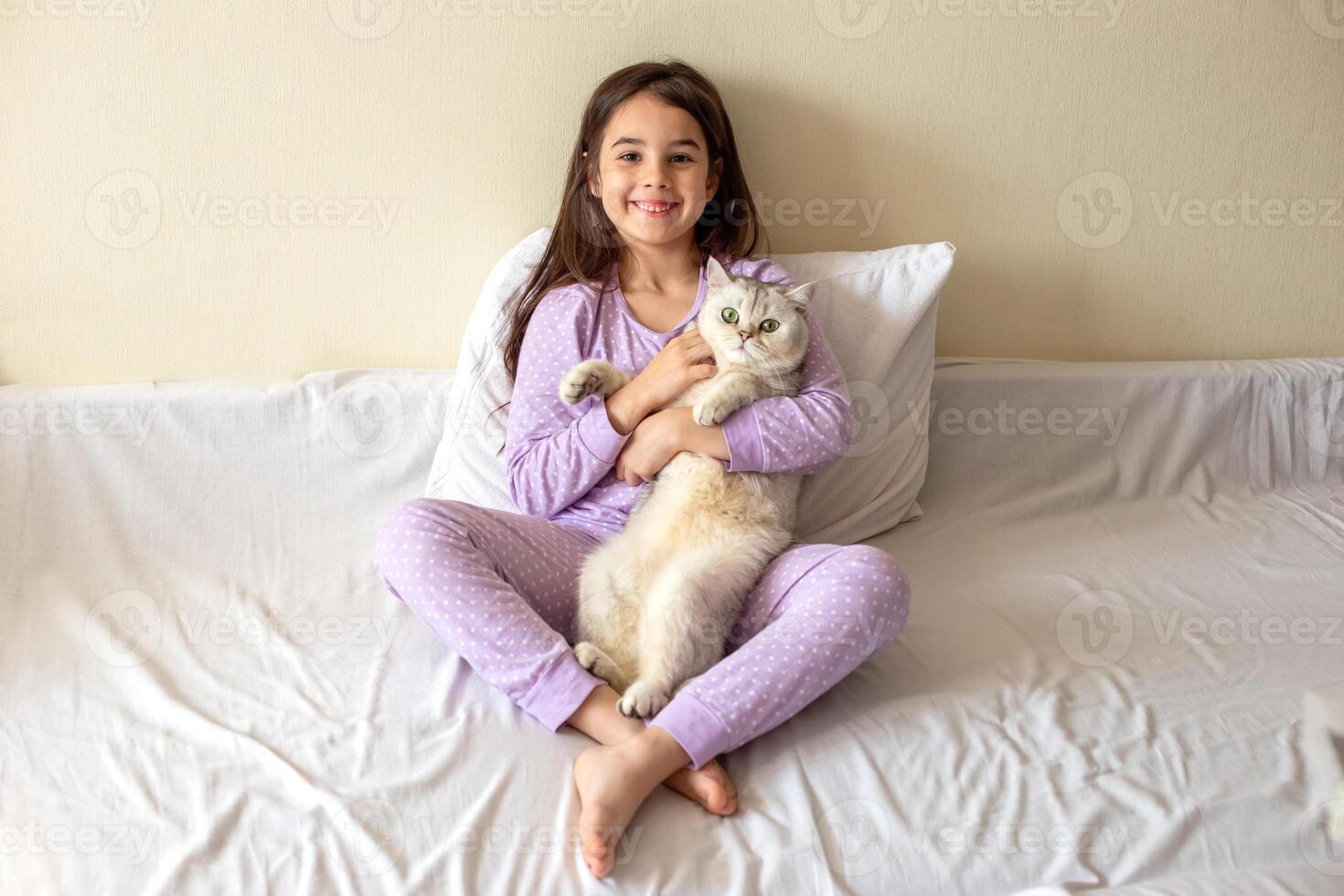 A happy little girl in purple pajamas, with her dark hair loose, sits hugging a white charming cat photo