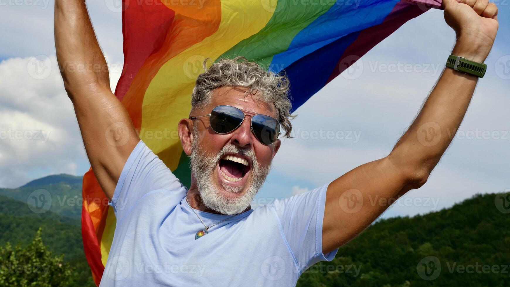 Portrait of a gray-haired elderly Caucasian man with a beard and sunglasses holding a rainbow LGBTQIA flag against a sky background, shouts in protest, Celebrates Pride Month Coming Out Day photo