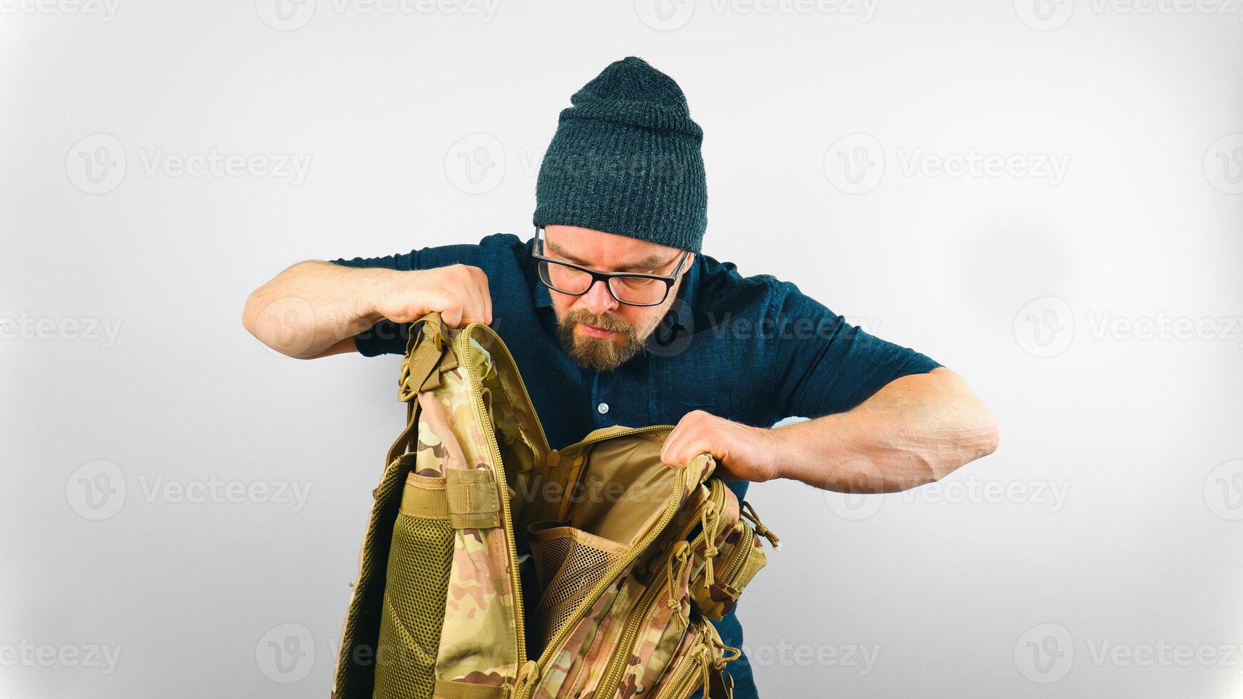 Portrait of handsome bearded man with beanie and glasses looking into camo military backpack, isolated on white background photo