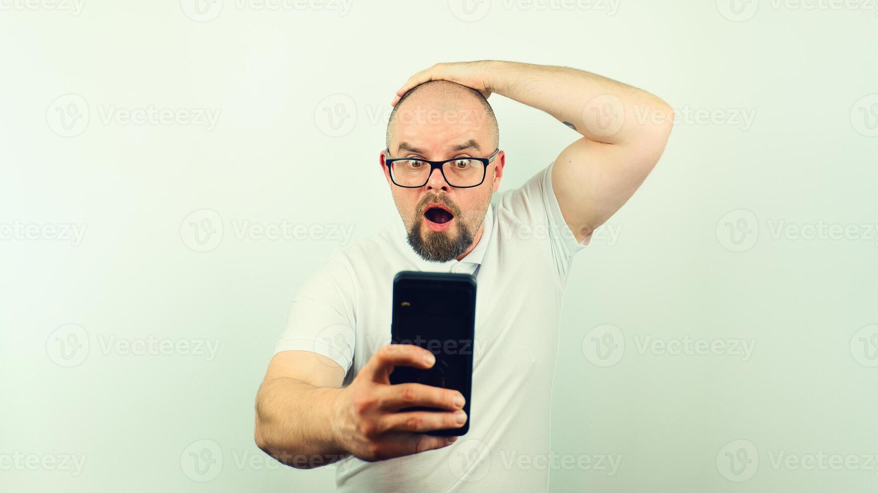 Portrait of surprised handsome bearded man with glasses and white shirt looking at mobile phone, facial expression, isolated on white background photo