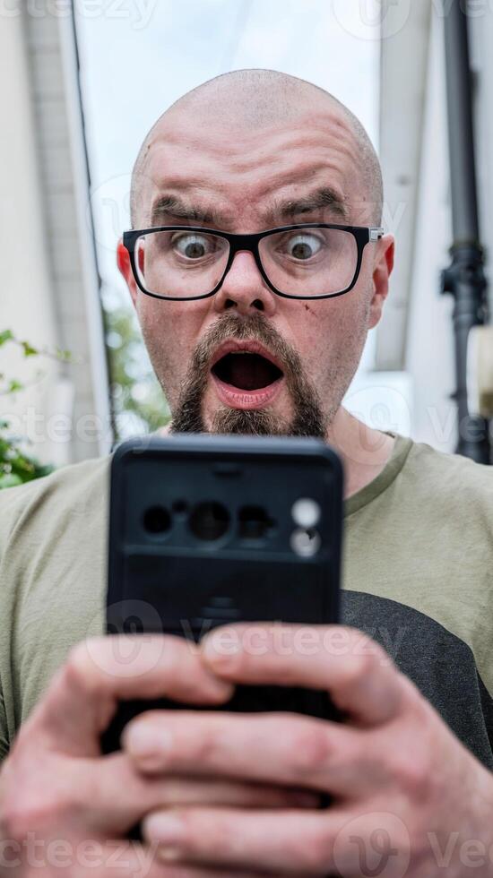 Portrait of surprised handsome bearded man with glasses looking at mobile phone, facial expression, people background photo