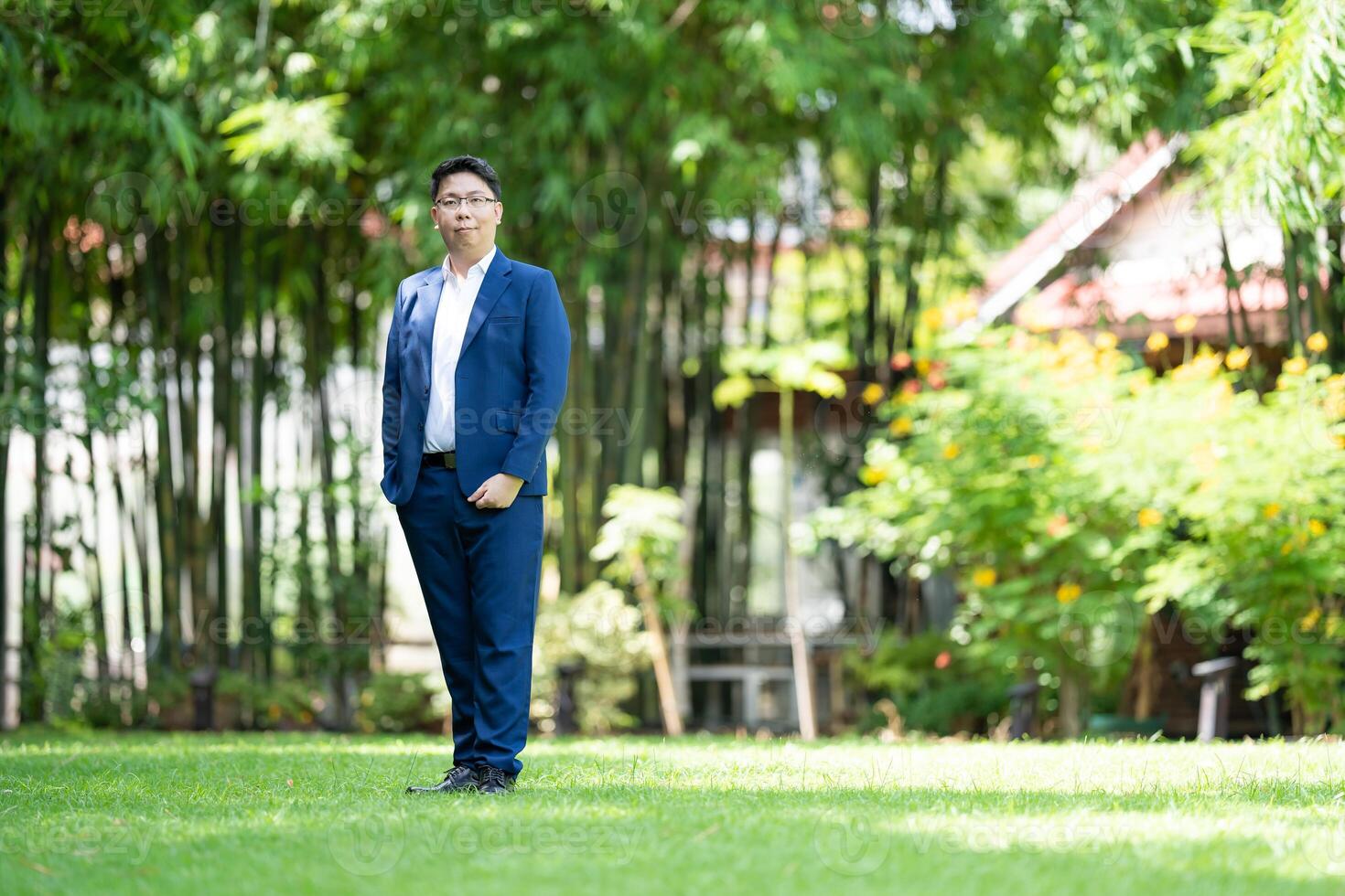 Asian handsome glasses man in a blue suit and the same color pants is looking at the camera, smart smiling and standing in the garden. photo