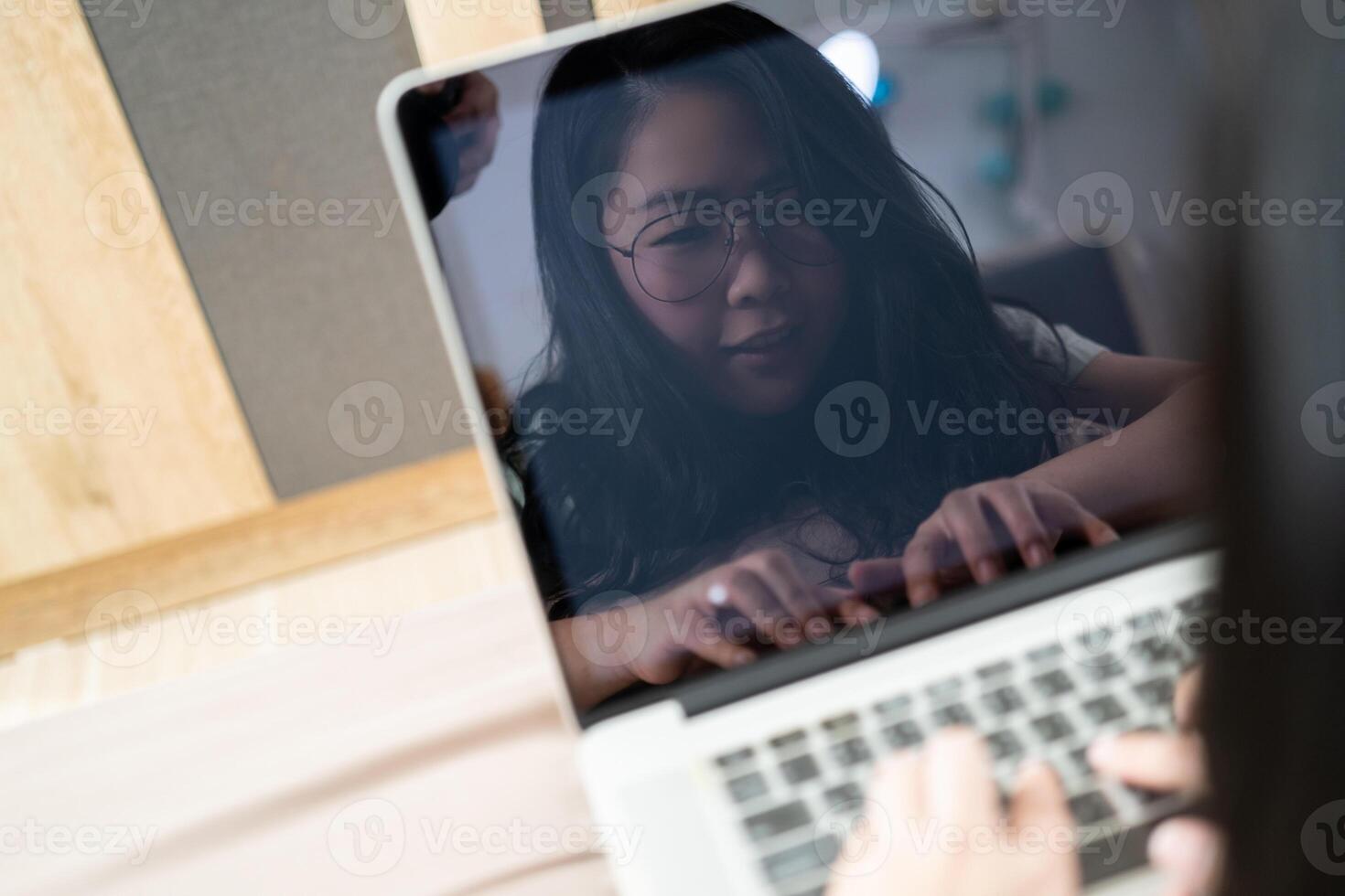 The reflection of Beautiful Asian woman from laptop screen. She plays laptop and lays on the bed in her bedroom. photo
