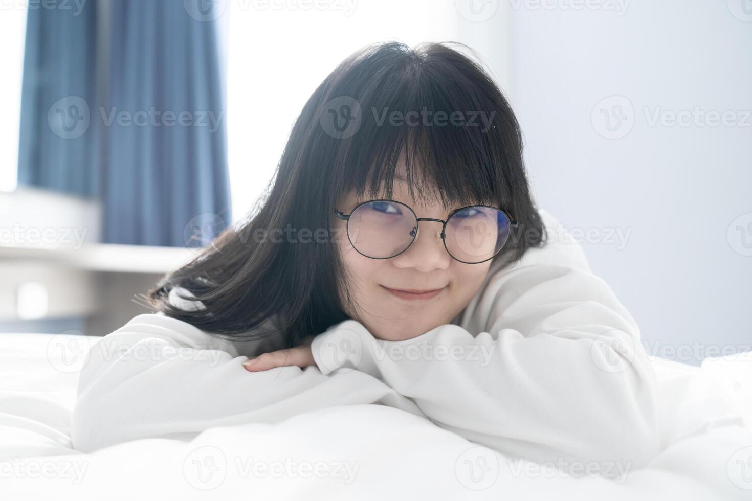 A sweet, cute Asian girl and her black glasses lays on the bed with happy and comfort feeling in front of window light in the morning. photo